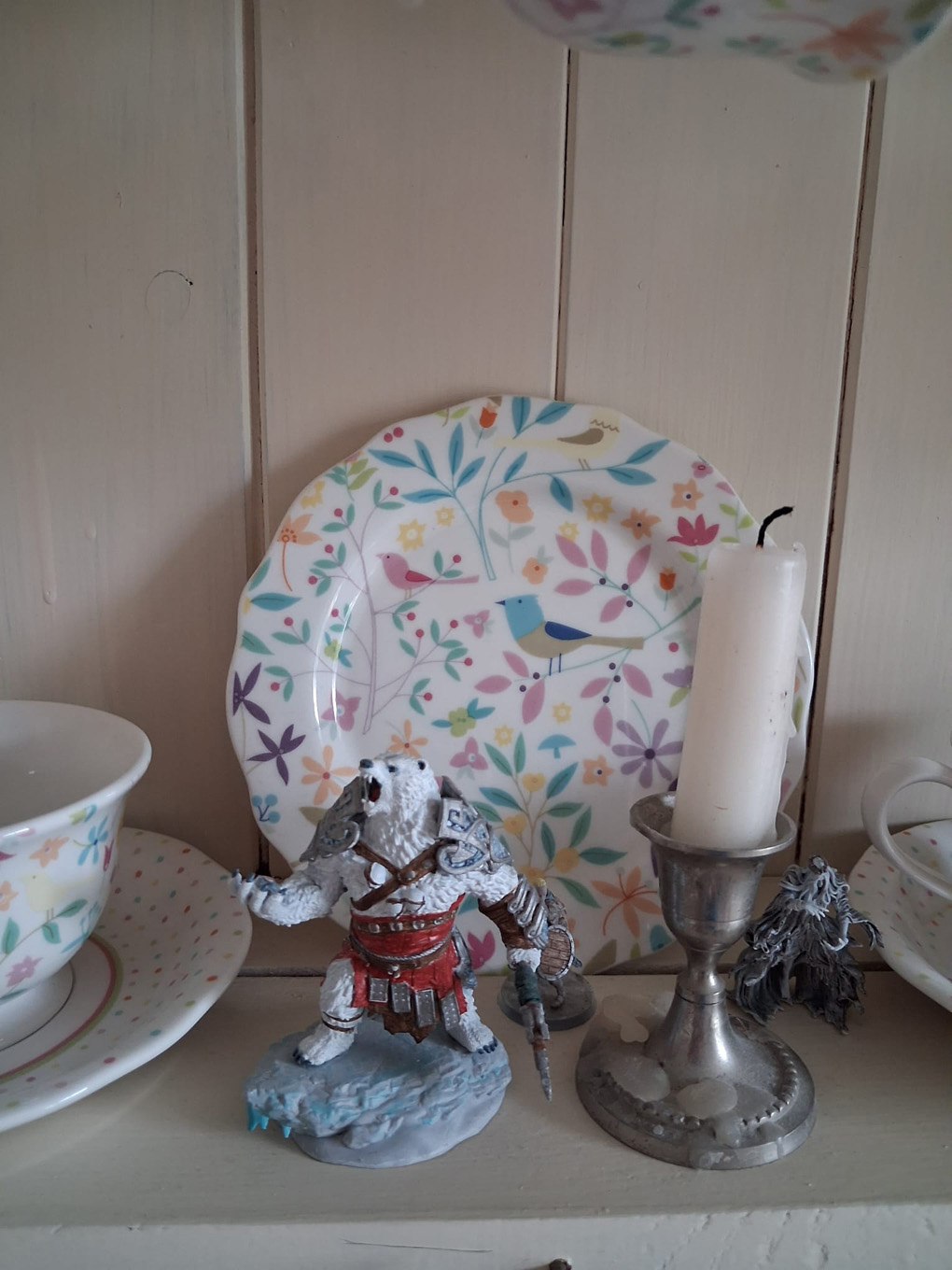 A DnD bear figurine mid roar, standing next to a candle, with a pretty floral plate displayed in the background.