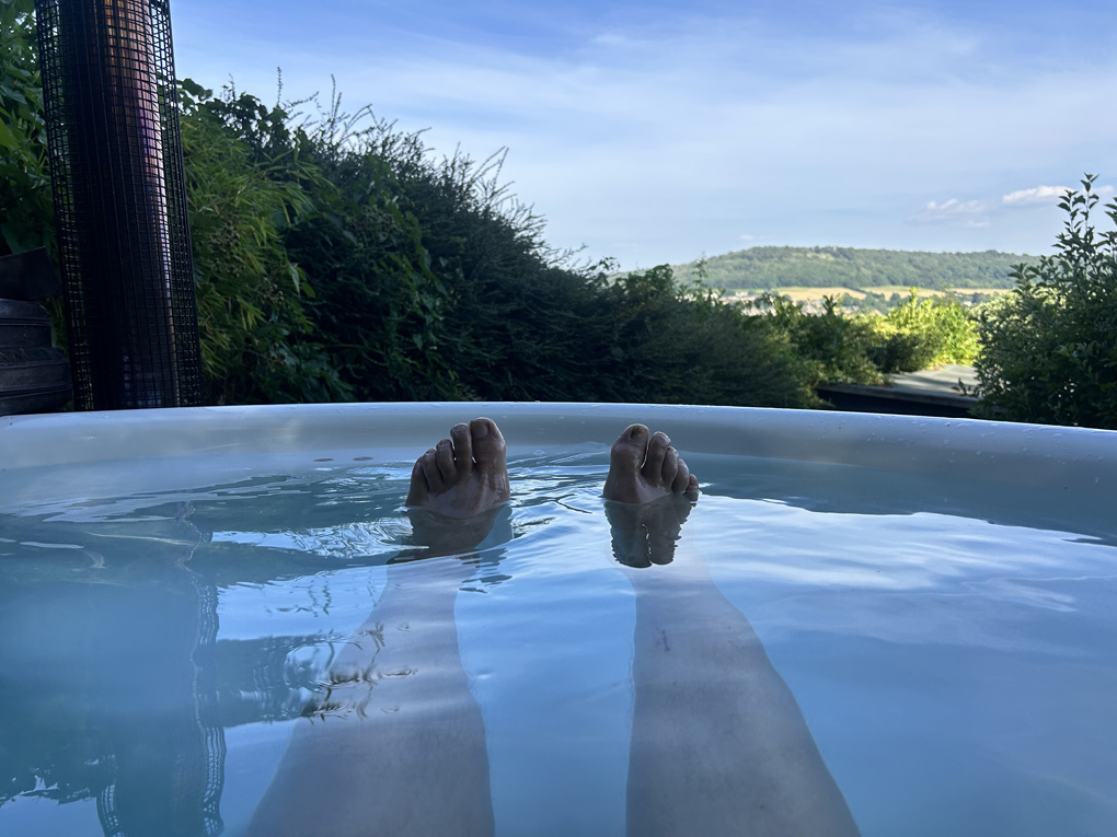 View from a hot tub with sunny hills in distance. Two legs floating on surface of the water from the person taking the photos and enjoying the coolness.