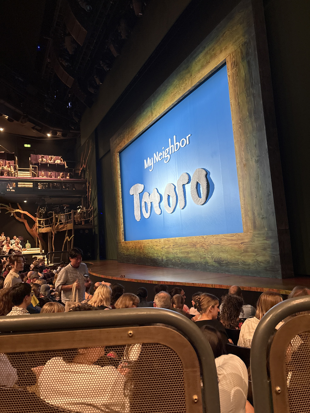 A seat view inside a theatre. The curtain in front of the stage is light blue with a green-ish orange wooden style frame around it. On the curtain are the words “My Neighbor Totoro”.