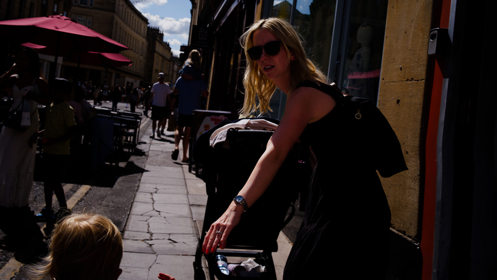Woman in a black sundress pushing a stroller reaches back for her toddler’s hand. Between the toddler and the mother is a branching crack in the sidewalk