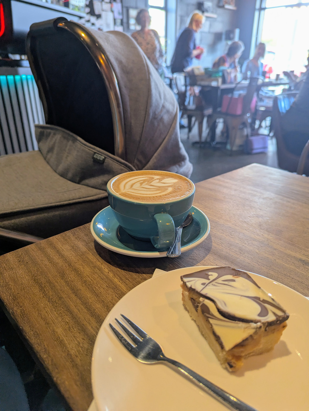 In the foreground, a cup of coffee and a cake against a background of a parked pram in a busy coffee shop