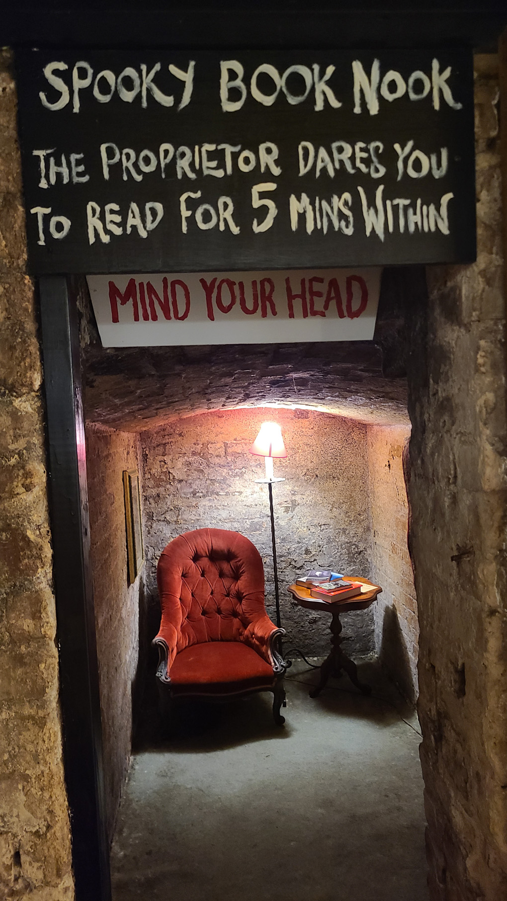 A red chair with a lamp to the right, in a brick alcove. A sign above reads - Spooky book nook. The proprietor dares you to read for 5 minutes within.