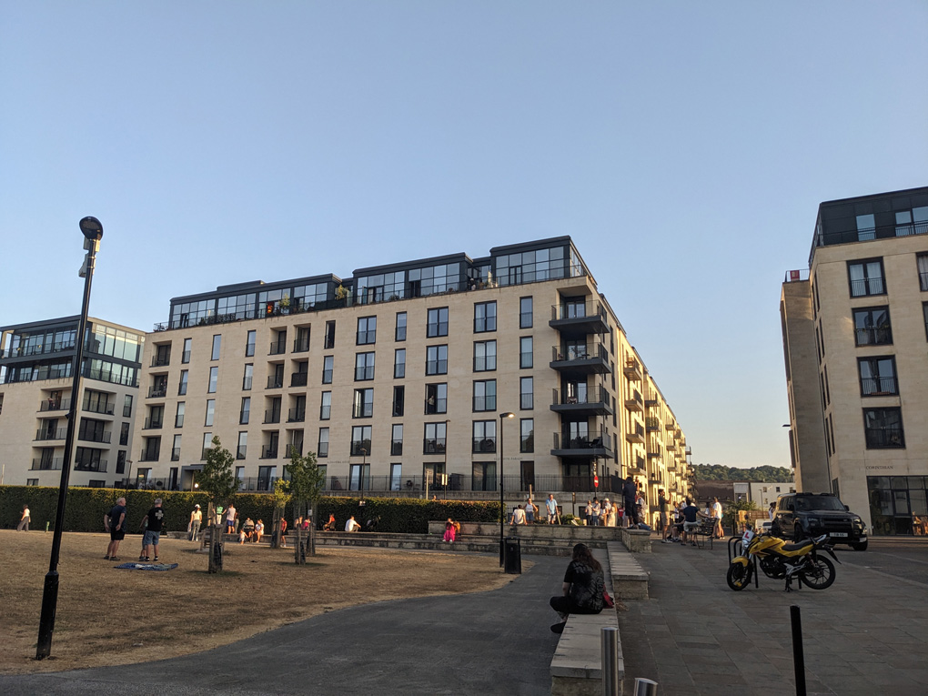 A park surrounded by beige apartment blocks with yellow grass and people stood or sat around looking at the sun