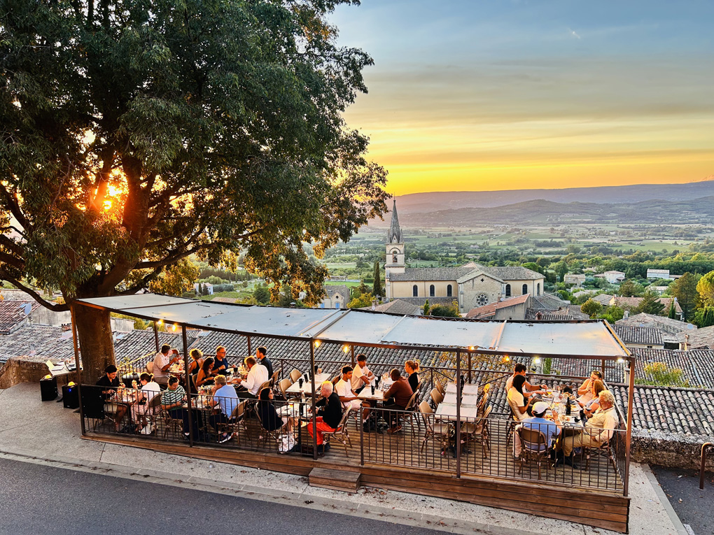 A wide, vibrant photograph taken during sunset shows a group of people dining outdoors on an elevated wooden terrace with a dark metal railing and an awning overhead. The terrace overlooks a scenic European town and a vast, lush valley framed by distant mountains.