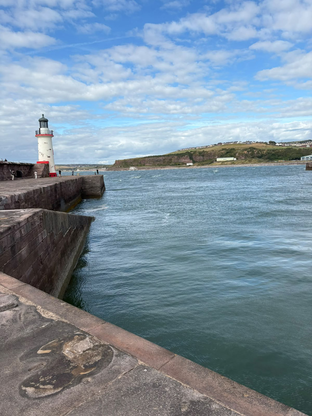 Clearly taken while standing on a large flat stone walkway, the main focus on the photo is the blue green sea. There is a predominantly white lighthouse with red highlights to the left side of the photo. There is a green/yellow land mass on the right side. The sky above is blue with thin cotton wool-like clouds