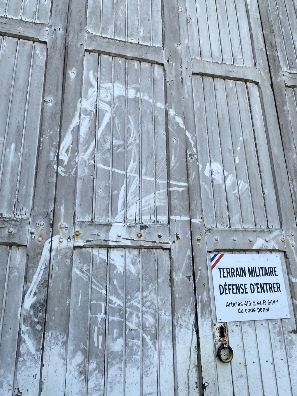 Dirty white wooden doors with a horse's outline drawn into the grime, beside a French 'Terrain Militaire' warning sign.