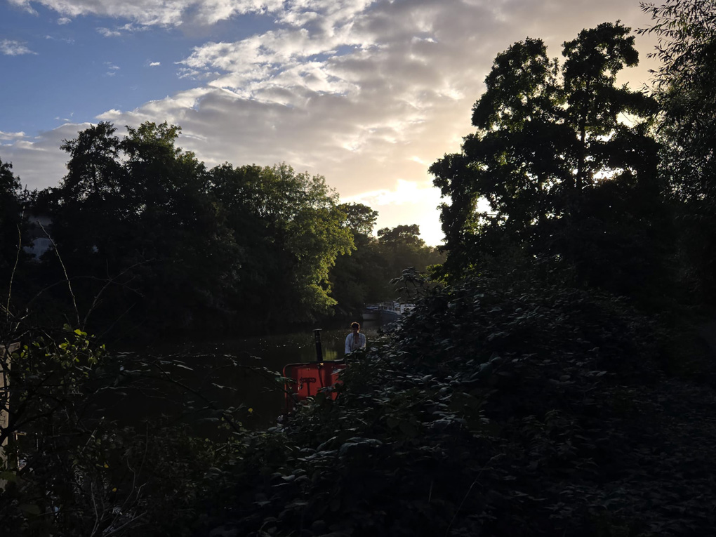 Most of the photo is greenery which lines the river. A canal boat is in view and the person operating it is bathed in the sunlight from a sun that is starting to set