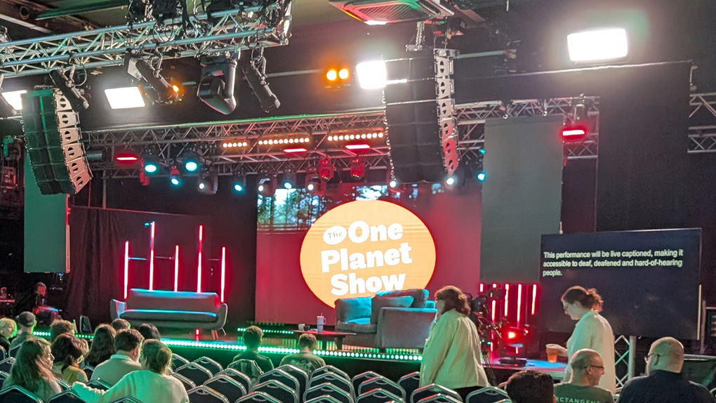 A stage set in readiness for a TV recording. There are two sofas on stage, one of which has a coffee table in front of it with a flask on it. There is a screen on the right explaining it will be live captioned. At the back of the set is the shows logo - the words The One Planet Show in white inside a large red circle. In the foreground we see seats, half of which are filled, with more audience members arriving.