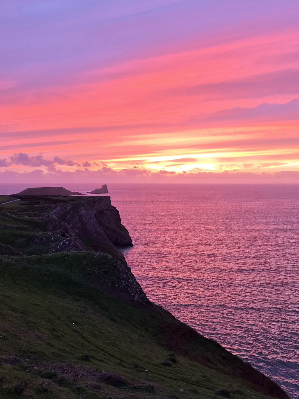 Pink sunset over pink coloured sea with dark green cliffs and a promontory of land stretching out into the sea towards the horizon.