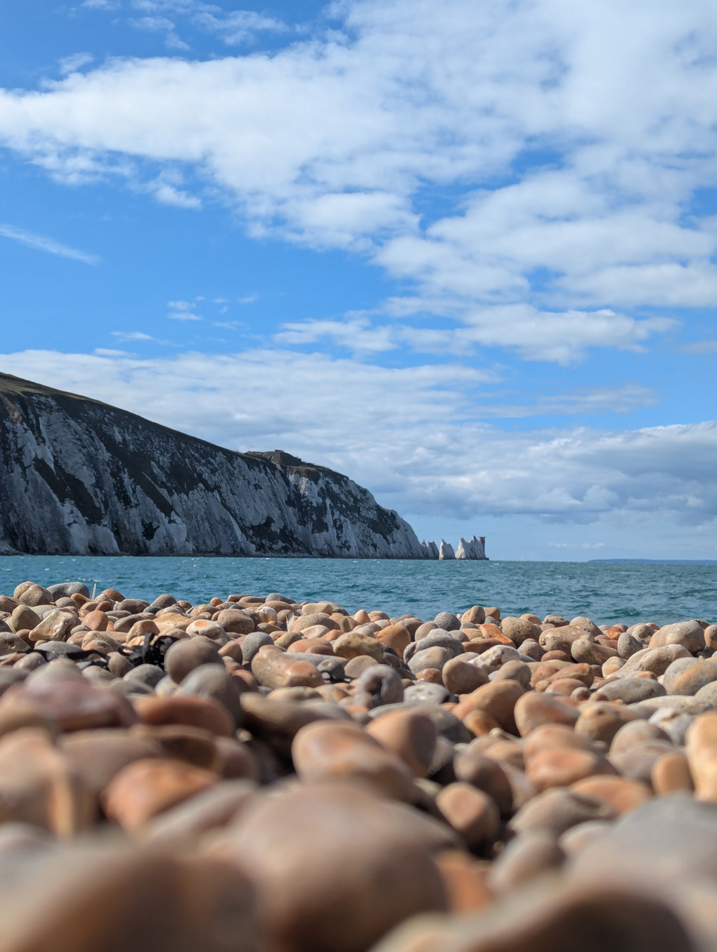 A view from a pebbled beach out across a clear expanse of blue sea to a stack of white cliffs and lighthouse in the far background