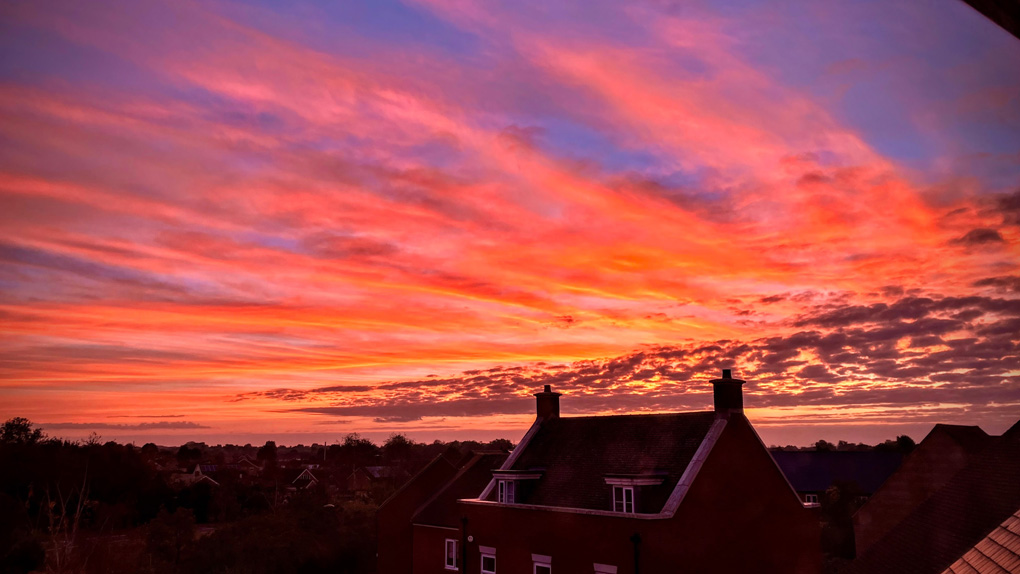 A dramatic morning sky that looks almost as though it is on fire, with dark, shadowy clouds in the distance and a neighbouring house silhouetted against the glowing orange horizon.
