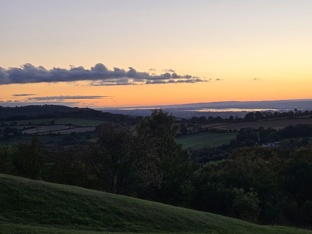View of green hills, river in the distance, orange tones in the sky as the sunsets and light bounces off the water