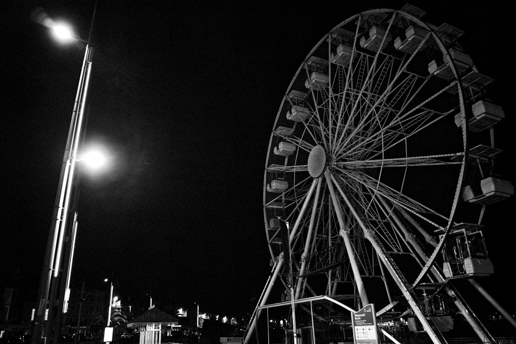 A motionless ferris wheel in the dead of night at the Weymouth seaside. It looks like a white skeleton against the night sky, looming overhead.