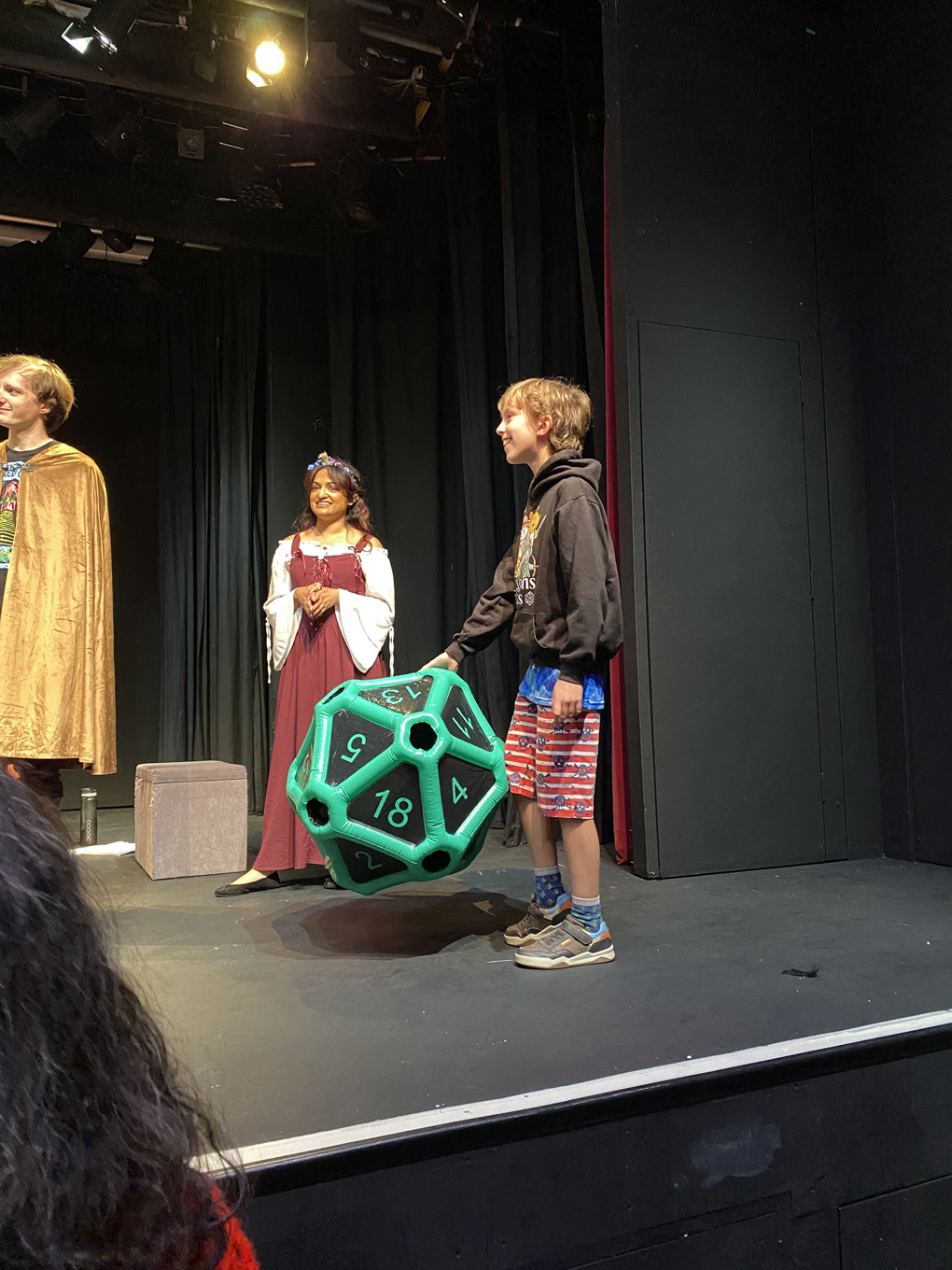 A boy smiles on stage while holding a giant inflatable twenty-sided die, with two costumed performers in fantasy dress behind him.