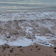 A pale sky just starting to colour with the sun going down, over the sea and a sandy and pebble beach.