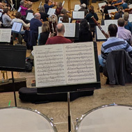 A view from the back of an orchestra, timpani in the foreground, and the orchestra spread out in a large hall.