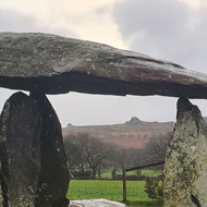 Ancient standing stone monument in green fields. A large flat stone lies upon three pointed standing stones in what appears to be impossible balance.