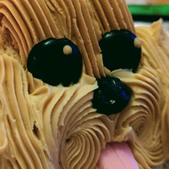 A long cake decorated with lots of swirled icing to look like a fluffy dog. Two jet black eyes and a tongue poke out from the face in the foreground