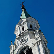 A blue sky, early morning, frames a picture of a large church spire beside which the metal fluted ring of the Winter Olympics cauldron - with its eternal flame - burns. Shot on a town square in the mountain town of Cortina, Italy.