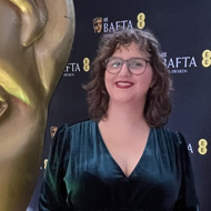 Me, a short brunette woman with glasses wearing a velvet green dress, standing on a red carpet next to a large statue of a BAFTA award