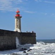 Photo taken from a shoreline. A lighthouse stands on a stone pier. A wave has crashed into a rock a few metres away, sending spray into the air.