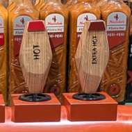 A scene from Nando's. A shelf of the normal sauces available, as normal - however in front of those is a dispenser, looking like the taps in a pub, with a nozzle for each flavour of source. It is a bright orange set of pumps dispensing Nando's sauce on-tap in flavours (Left to Right) - Lemon & Herb, Medium, Wild Herb, Garlic, Hot, Extra Hot. There is a drip tray under the whole thing.