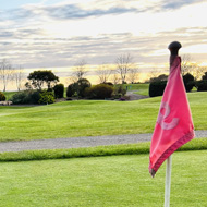 Sunrise at a golf club. A golf ball site beside a hole with the flag in, and in the background the sun rises and dapples the clouds in a mixture of red and orange.