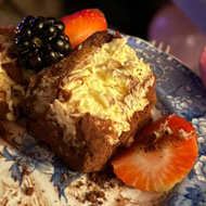 A slice of tiramisu lays on a blue and white plate with two slices of strawberry and a blackberry on top