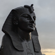 A large dark bronze sphinx on a plinth with the river Thames behind and high rise city buildings in the distance on a slightly grey early morning with the sun just starting to glow through on the horizon