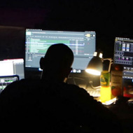 The main subject is a single figure on spotlighted on stage. In the foreground are technicians silhouetted by the lights from their many monitor screens.