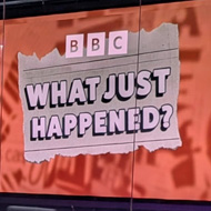 In the foreground, a long, purple and white desk set up for a panel show with five empty chairs and five microphones for the host/panel. The set itself is mainly purple. Behind the desk is a window which looks out over a foyer space, but at the back of the foyer is a large digital display with the show's logo - an orange design and the words 