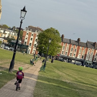 Two children ride away from the camera along a path across Blackheath, wearing helmets, heading toward a Victorian church spire. A park bench and lamp post sit to the left, with London terraces in the background under an evening sky.