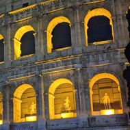 The Rome Colosseum at night, with each recessed arch lit in dark orange light illuminating the statues in each, as they would have been in place in ancient rome
