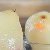 Two glasses with white liquid. On top of each glass is a translucent looking fruit, peeled and slightly damp. Held in place by a plastic stick with a heart shaped end. In the background are sunny mountains covered in snow.