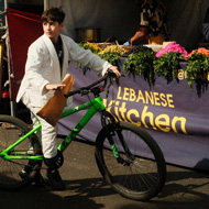 a kid in a white karate gi, on a green bike, is stopped in front of a colorful lebanese food stall in broadway market. The light frames and highlights his face with warmth, contrasting the shadows around him.