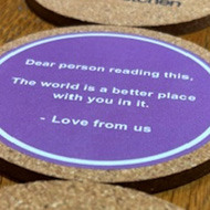 A line of beer mats with one showing the underside that shows the message 'Dear person reading this, the world is a better place with you in it' - Love from us