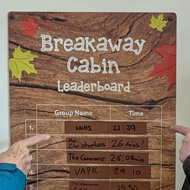 Four family members standing around an escape room leaderboard of escape times, pointing at their team at the top.