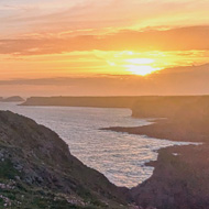A few figures in silhouette walking along a cliff top with the sea stretching before them and a glowing sunset in the background