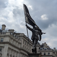 A statue surrounded by important buildings, embassies, government and royalty. One of many statues in the area. The person in this statue is carrying a flag which has blown back to cover their face. They are marching forward off the plinth.