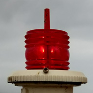 Very rusty bracket suporting a red navigation lamp, with seascape behind.