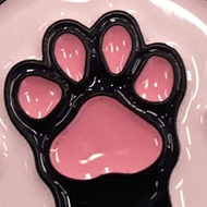Round pink enamel pin featuring a black cat paw with pink toe beans at the centre, flanked by two red hearts. The text 