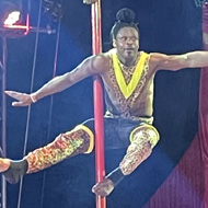 Four brightly dressed circus acrobats balance vertically on a tall red pole, one above the other, with their arms stretched out under colourful stage lights.