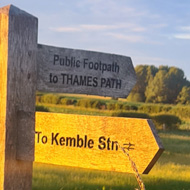 Wooden footpath sign at a field stile pointing to Kemble Station and the Thames Path with green wild foliage and cow parsley bathed in golden light