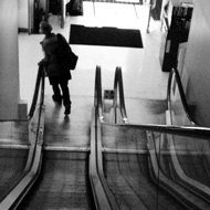 A black and white image shot from the top of the escalator in the Podium, looking down from the library landing. a man is tilting to his lift as he fast walks around the corner from the bottom of the escalator to the doors to the street. strong contrast from the black of the escalator and the man's silhouette with the light glinting off of edges in the late afternoon.