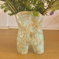 A selection of brightly coloured and interestingly textured flowers in a vase shaped like a woman's torso in a swimming costume. On a brown kitchen counter with white tiles behind.