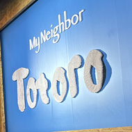 A seat view inside a theatre. The curtain in front of the stage is light blue with a green-ish orange wooden style frame around it. On the curtain are the words “My Neighbor Totoro”.