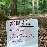 A 'Longest Putt' competition sign for on a disc golf course, staked into fallen leaves, with a disc golf basket visible among the trees behind it.