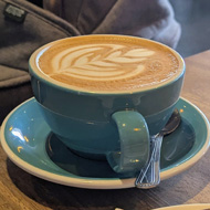 In the foreground, a cup of coffee and a cake against a background of a parked pram in a busy coffee shop