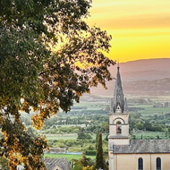 A wide, vibrant photograph taken during sunset shows a group of people dining outdoors on an elevated wooden terrace with a dark metal railing and an awning overhead. The terrace overlooks a scenic European town and a vast, lush valley framed by distant mountains.