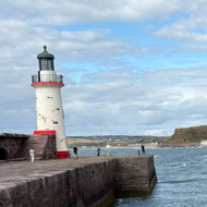 Clearly taken while standing on a large flat stone walkway, the main focus on the photo is the blue green sea. There is a predominantly white lighthouse with red highlights to the left side of the photo. There is a green/yellow land mass on the right side. The sky above is blue with thin cotton wool-like clouds